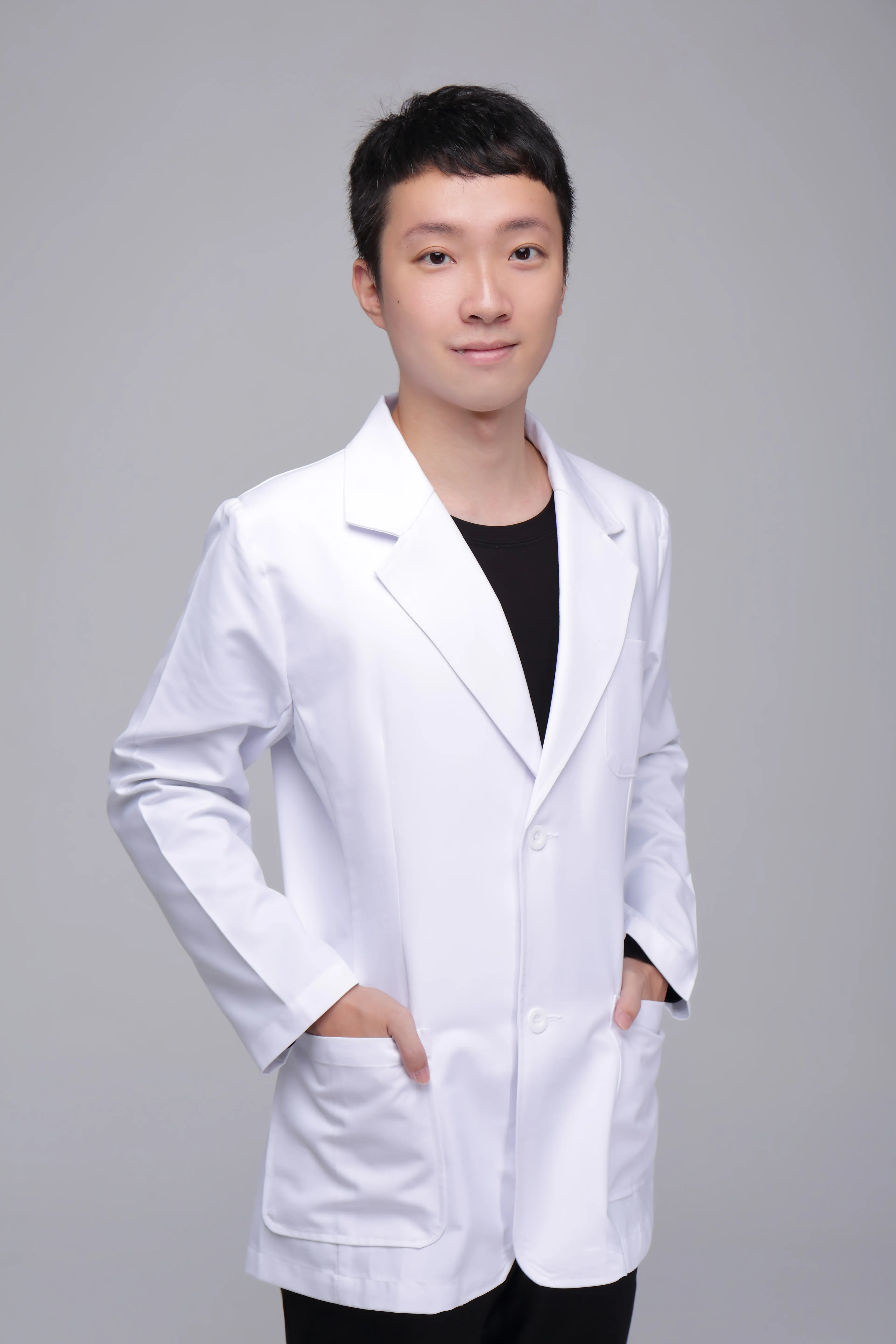 Person in a white lab coat smiling with hands in pockets, against a plain gray background.